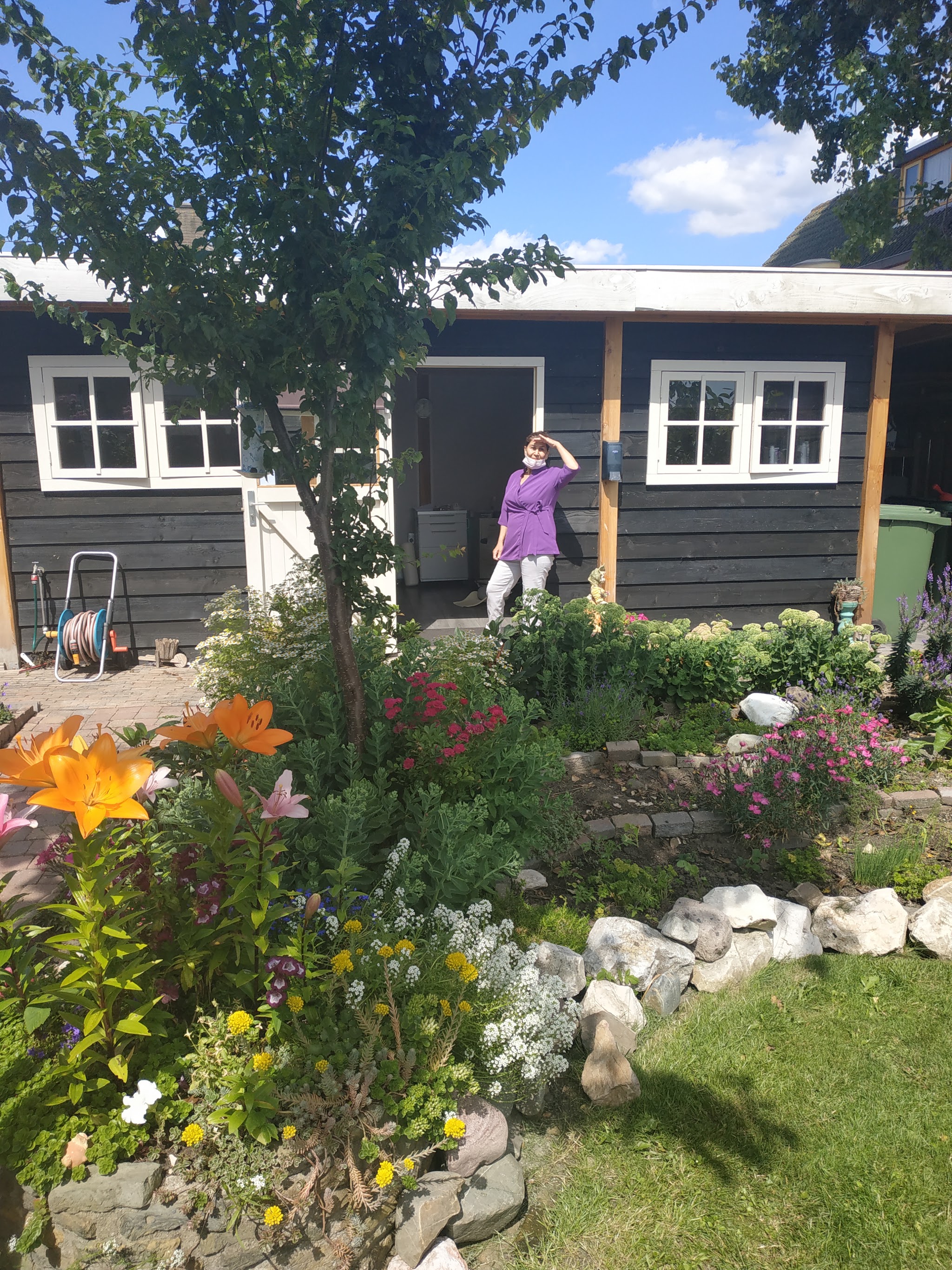 De tuin met bloemen en de donkerblauwe houten praktijk, Aki staat in de deuropening